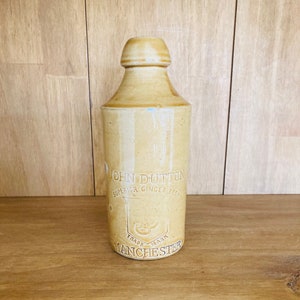 Op de afbeelding: Een vintage gemberbierfles van steengoed in een beige kleur. De fles heeft een afgeronde bovenkant en een cilindrisch lichaam. De tekst "JOHN DUTTON SUPERIOR GINGER BEER TRADE MARK MANCHESTER" is op de voorkant in reliëf aangebracht.