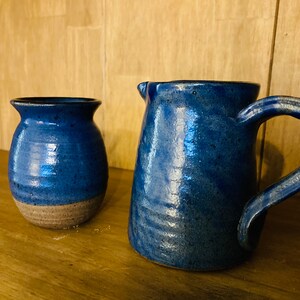 May include: A blue ceramic jug and vase set. The jug has a handle and spout, while the vase is a smaller, rounded shape. Both pieces have a speckled blue glaze with a natural, earthy aesthetic.