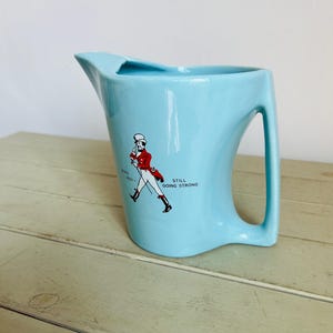 Puede incluir: Jarra de cer&aacute;mica azul claro con asa y boquilla. La jarra presenta un gr&aacute;fico de un hombre con abrigo rojo y el texto "BORN 1820 - STILL GOING STRONG".