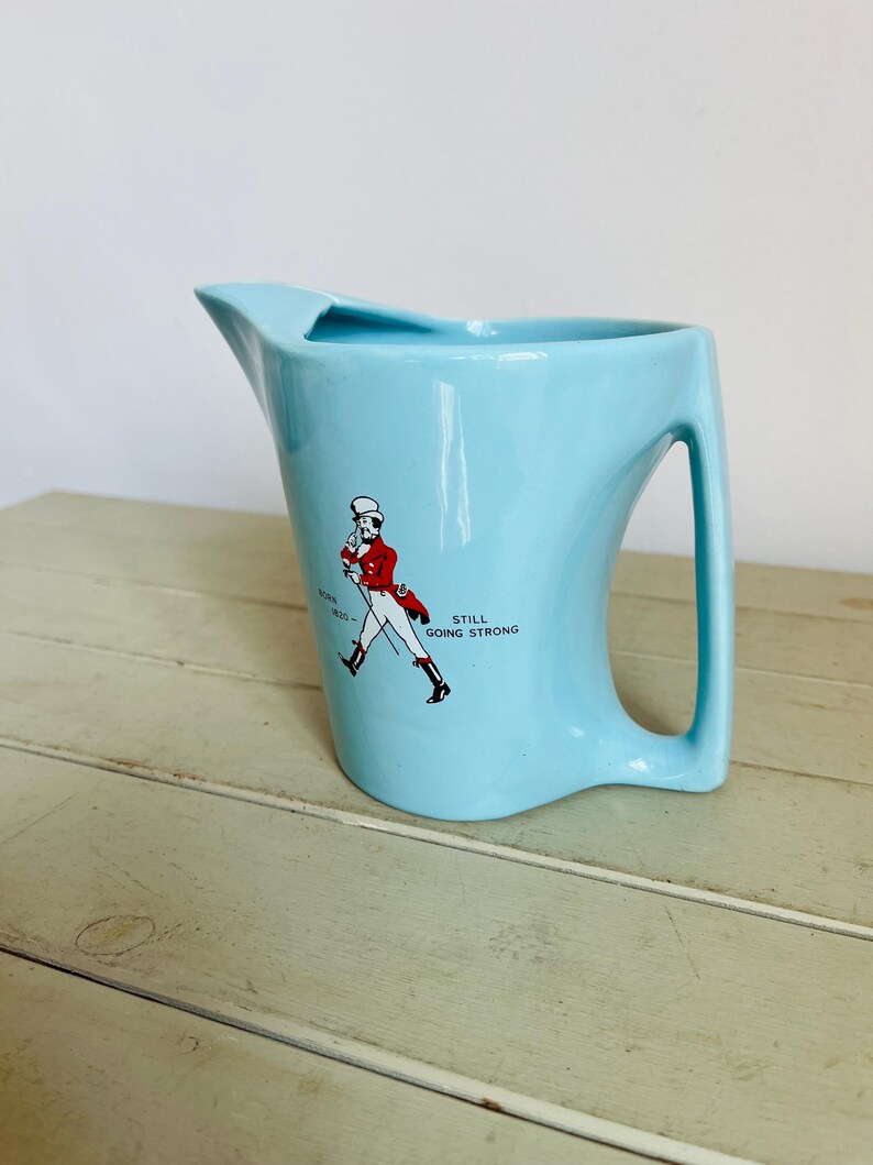 Puede incluir: Jarra de cer&aacute;mica azul claro con asa y boquilla. La jarra presenta un gr&aacute;fico de un hombre con abrigo rojo y el texto "BORN 1820 - STILL GOING STRONG".