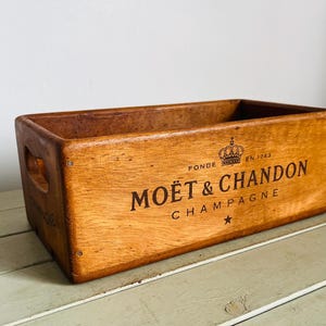 Puede incluir: Una caja rectangular de madera con un acabado marrón medio y asas recortadas en los laterales. El panel frontal presenta un texto impreso en negro que dice MOËT & CHANDON CHAMPAGNE, acompañado de un icono de corona, el texto FONDE EN 1743 y una estrella.