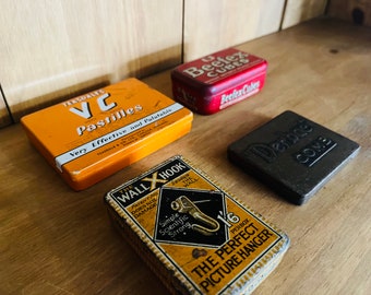 Pode incluir: Quatro caixas de lata vintage com vários designs. As caixas são amarelas, vermelhas, pretas e amarelas com texto preto. O texto nas caixas diz "Tensdale VC Pastilles Very Effective and Palatable", "Beetex Cubes", "Derdae Come", e "The Wall X Hook The Perfect Picture Hanger".