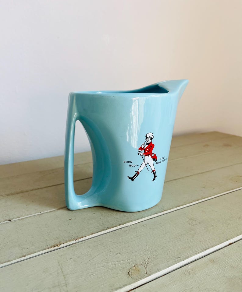 Puede incluir: Jarra de cer&aacute;mica azul claro con un dise&ntilde;o de asa &uacute;nico. La jarra presenta un gr&aacute;fico de un hombre con abrigo rojo y el texto "BORN 1820 - STILL GOING STRONG". La jarra tiene un pico para verter f&aacute;cilmente.