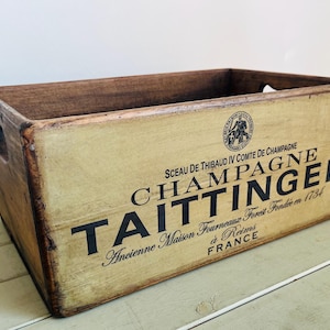 Può includere: Una cassa in legno rettangolare con un pannello frontale color crema dall'aspetto vissuto che riporta la scritta nera Champagne Taittinger, Sceau de Thibaud IV Comte de Champagne, Ancienne Maison Fourneaux Forest Fondée en 1734 à Reims France.