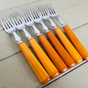 Puede incluir: Seis tenedores vintage con púas de acero inoxidable y un diseño decorativo en la base. Los mangos son de un color ámbar-naranja cálido. Los tenedores están dispuestos diagonalmente sobre una superficie de madera clara.
