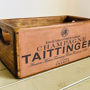 May include: A rectangular wooden crate with cut-out handles on the sides. The front panel features a light brown surface with black text reading Champagne Taittinger, Sceau de Thibaud IV Comte de Champagne, Ancienne Maison Fourneaux, and France.
