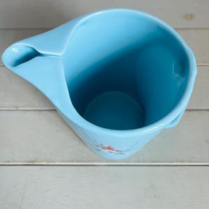 Puede incluir: Una jarra de cer&aacute;mica azul claro con pico y asa. La jarra tiene un acabado liso y brillante y una forma ligeramente acampanada. Un peque&ntilde;o dise&ntilde;o floral es visible cerca de la base. La jarra est&aacute; vac&iacute;a y parece limpia.