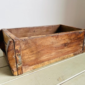 Puede incluir: Una caja rectangular de madera con un acabado envejecido. La caja está construida con gruesas tablas de madera y soportes metálicos en las esquinas. La madera tiene un tono marrón cálido con vetas visibles y signos de desgaste.