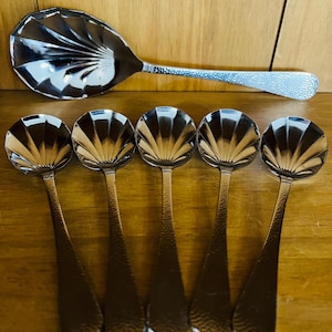 Può includere: Un set di sei cucchiai da portata color argento. Il cucchiaio grande ha una coppetta a forma di ventaglio e un manico strutturato. Sotto ci sono cinque cucchiai più piccoli con coppette a forma di conchiglia e manici strutturati. I cucchiai sono su una superficie di legno.