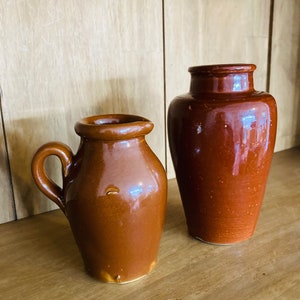 May include: Two brown ceramic pottery pieces, a small jug with a handle and a larger vase. Both pieces have a glossy finish.