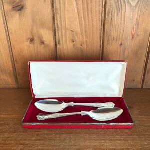 Op de afbeelding: Twee zilveren lepels in een rode fluwelen doos. De lepels hebben sierlijke handvatten en zitten in een presentatiedoos.