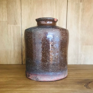 Puede incluir: Un tarro de cerámica marrón oscuro con una boca ancha y un cuerpo ligeramente redondeado. El tarro tiene una superficie rugosa y texturizada y un borde ligeramente irregular.