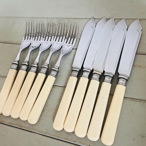 Puede incluir: Un conjunto de cubiertos antiguos, que incluye tenedores y cuchillos. Los tenedores tienen cuatro púas y los cuchillos tienen una hoja curva. Ambos tienen mangos color crema y detalles plateados.