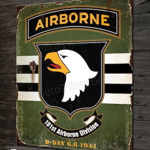 Puede incluir: Un letrero de metal de estilo vintage con fondo verde y un emblema de águila en blanco y negro. El letrero dice "AIRBORNE" en letras amarillas y "101st Airborne Division" en letras blancas. El letrero también incluye el texto "D-DAY 6.6.1944" en letras blancas.