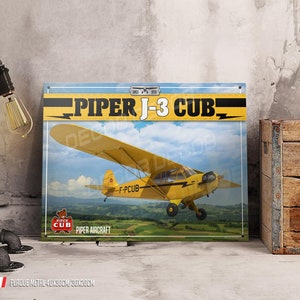 Könnte beinhalten: Ein gelbes Piper J-3 Cub Flugzeug mit dem Kennzeichen F-PCUB fliegt über ein grünes Feld. Das Metallschild zeigt "Piper J-3 Cub" und "Piper Aircraft".