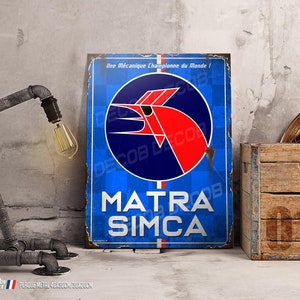 Puede incluir: Una placa de metal azul y roja con el texto "Une Mécanique Championne du Monde!" y "MATRA SIMCA". La placa presenta un logotipo de pájaro rojo estilizado sobre un círculo azul con un fondo a cuadros.
