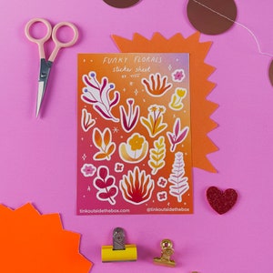 Op de afbeelding: Een vel stickers met verschillende kleurrijke bloemenontwerpen. Het vel is getiteld "Funky Florals Sticker Sheet by Tian" en heeft een verloopachtergrond in roze, oranje en geel. Het vel is omringd door knutselbenodigdheden, waaronder een schaar, paperclips en een hartvormige versiering.