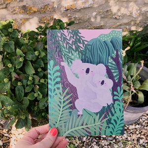 May include: A greeting card featuring a pink and green illustration of two koalas in a jungle setting. The koalas are light purple and have black eyes and noses. The background is a lush green jungle with pink and purple accents.