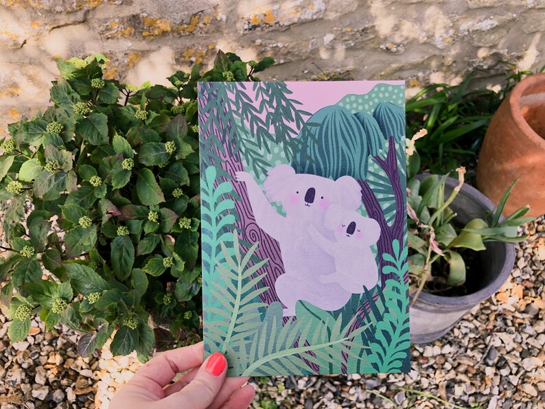 May include: A greeting card featuring a pink and green illustration of two koalas in a jungle setting. The koalas are light purple and have black eyes and noses. The background is a lush green jungle with pink and purple accents.
