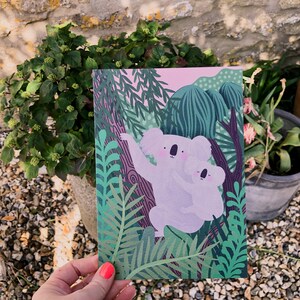 May include: A pink and green illustrated card featuring two koalas hugging in a jungle setting. The koalas are white with pink noses and black eyes. The jungle is filled with green leaves and vines.