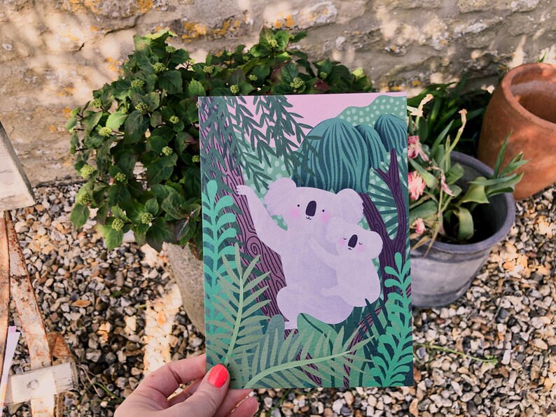 May include: A pink and green illustrated card featuring two koalas hugging in a jungle setting. The koalas are white with pink noses and black eyes. The jungle is filled with green leaves and vines.