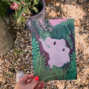May include: A pink and green illustrated card featuring two koalas hugging in a lush green forest setting.
