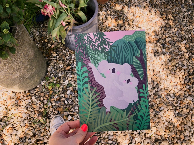 May include: A pink and green illustrated card featuring two koalas hugging in a lush green forest setting.