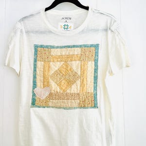 以下が含まれることがあります： J.CrewのサイズMのタグが付いた白い半袖Tシャツ。フロントには黄色とティールカラーの正方形のパッチワークデザインが施されており、左下隅には小さなハートのアップリケが付いています。袖には繊細なレースのトリムがあしらわれています。
