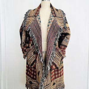 May include: A long, open-front cardigan in a patchwork design. The coat features a mix of patterns in shades of brown, burgundy, and tan, with fringed edges. The garment is displayed on a mannequin, showcasing its length and style.