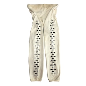 May include: Cream-colored sweatpants with black bat-shaped graphics down the sides. The sweatpants have a drawstring waistband and elastic cuffs. The text "Powell Peralta" is printed on the bottom of each leg.