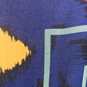 May include: Close-up of a blue fabric with a colorful abstract pattern. The pattern includes black, yellow, pink, and purple brushstrokes. The fabric has a textured weave.