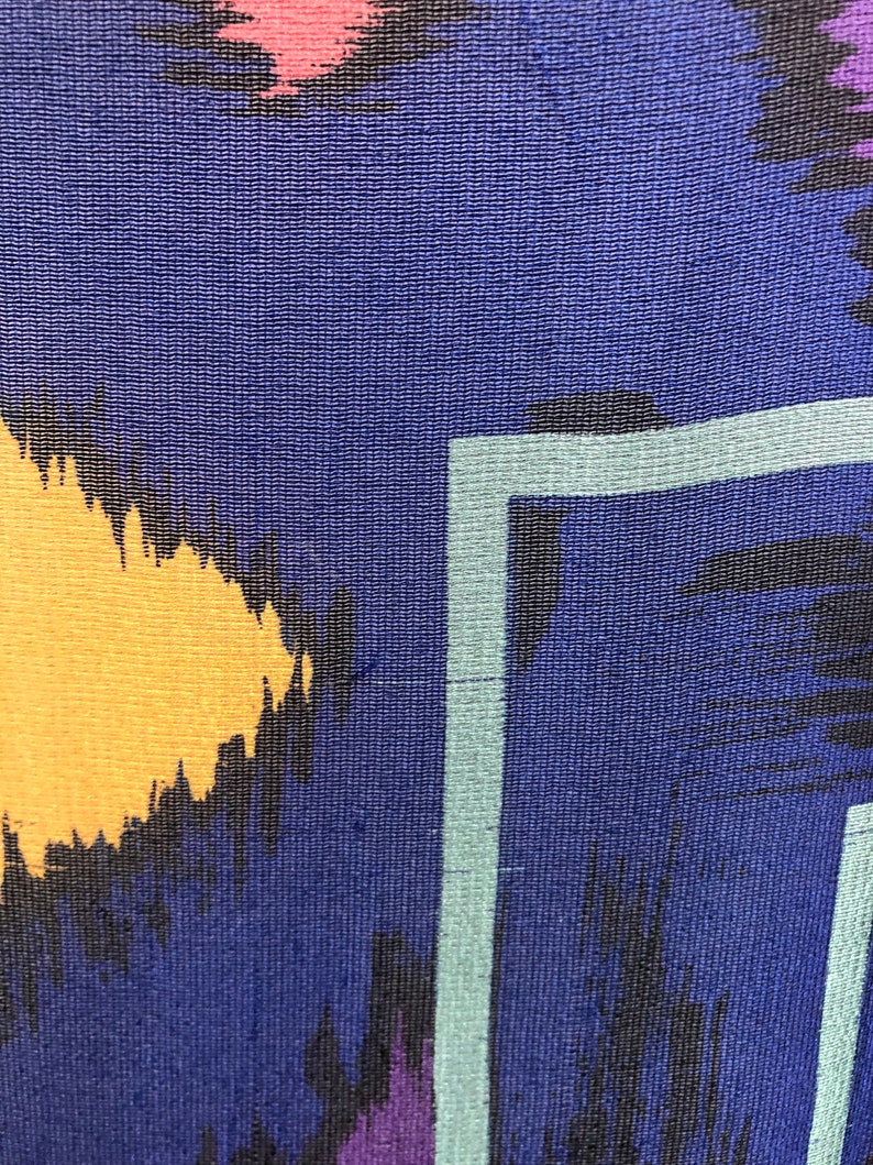 May include: Close-up of a blue fabric with a colorful abstract pattern. The pattern includes black, yellow, pink, and purple brushstrokes. The fabric has a textured weave.