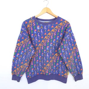 May include: A vintage blue sweatshirt with a repeating pattern of the word "AUSTRALIA" in various colors and shapes. The long-sleeved top has a crew neck and ribbed cuffs and hem. The sweatshirt is hanging on a wooden hanger.