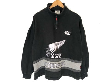 Puede incluir: Sudadera negra de manga larga con un logotipo blanco de los All Blacks de Nueva Zelanda y una franja plateada y gris en la parte inferior. El logotipo presenta un helecho plateado y el texto "NEW ZEALAND ALL BLACKS". La franja tiene la palabra "AUTHENTIC" en blanco.