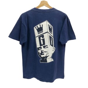 May include: Navy blue t-shirt with a graphic on the back featuring a stylized face with a crown and the letter "G" in a box. The design is in white, contrasting against the dark blue fabric. The shirt is hanging on a wooden hanger.