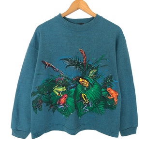 May include: Teal-colored sweatshirt featuring a vibrant jungle scene with various colorful frogs. The graphic includes red, yellow, and green frogs amidst lush green foliage. The long-sleeved top is hanging on a wooden hanger.