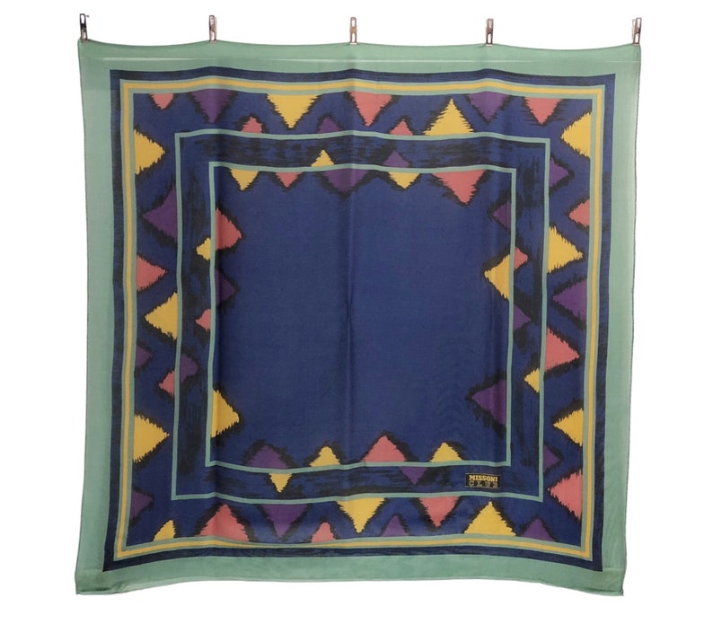 May include: A blue and green patterned silk scarf with a geometric design. The scarf has a border of yellow, pink, and purple triangles. The center of the scarf is a solid blue color. The scarf has the text "MISSOMA" in the bottom right corner.