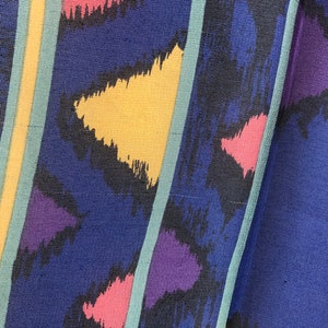 May include: A close-up of a blue fabric with a colorful abstract pattern. The pattern features yellow, pink, purple, and black brushstrokes. The fabric has a textured surface.