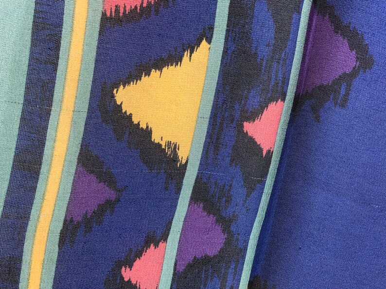 May include: A close-up of a blue fabric with a colorful abstract pattern. The pattern features yellow, pink, purple, and black brushstrokes. The fabric has a textured surface.