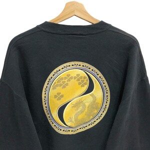 Puede incluir: Sudadera negra con un gran s&iacute;mbolo yin-yang dorado que presenta un drag&oacute;n y un dise&ntilde;o floral. El s&iacute;mbolo est&aacute; rodeado por un borde negro y morado con caracteres repetidos.