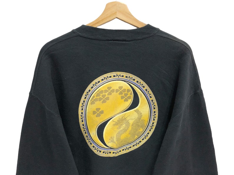 Puede incluir: Sudadera negra con un gran s&iacute;mbolo yin-yang dorado que presenta un drag&oacute;n y un dise&ntilde;o floral. El s&iacute;mbolo est&aacute; rodeado por un borde negro y morado con caracteres repetidos.