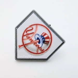 May include: A home plate-shaped pin featuring the New York Yankees logo. The logo includes the word "Yankees" in red script, a baseball, and a patriotic design in red, white, and blue. The pin has a gray border and the text "DecoPac/MLB".