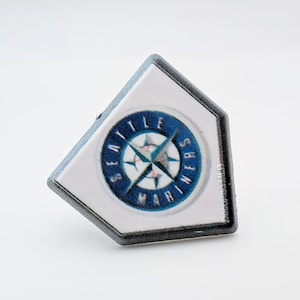 May include: A white, home plate-shaped pin featuring the Seattle Mariners logo. The logo is a blue and white compass rose design with the words "SEATTLE MARINERS" in a circular pattern. The pin has a black border.