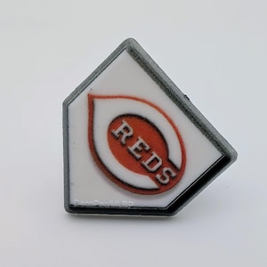 May include: A white, diamond-shaped pin with a black border. The pin features the Cincinnati Reds logo, with the word "REDS" in red letters inside a red oval.
