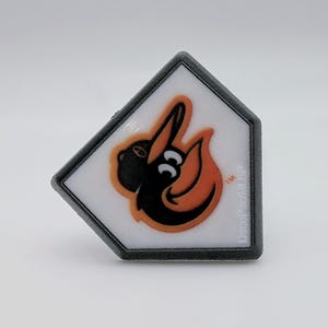 May include: A baseball-themed magnet featuring the Baltimore Orioles logo. The logo is a stylized bird in black and orange, set against a white background within a home plate-shaped frame. The frame is outlined in black.
