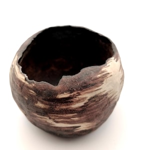 May include: A handcrafted ceramic bowl with a textured surface. The bowl has a dark brown and cream colour scheme and an irregular shape. The interior is dark, contrasting with the lighter exterior. The bowl is approximately 10 cm in diameter.