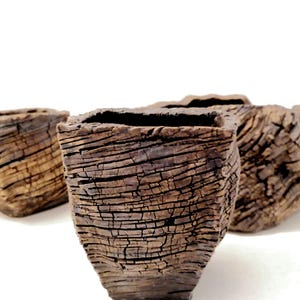 May include: Three square-shaped ceramic vessels with a dark brown, deeply textured exterior that mimics the appearance of weathered tree bark. Each container features a rough, cracked surface pattern and a hollow, open top with a square rim.