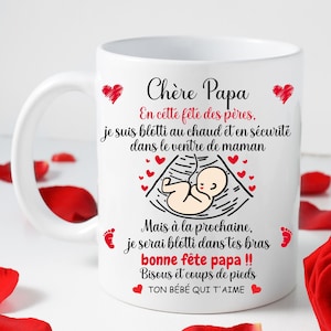 Peut inclure: Mug blanc avec une anse, orné de texte et d'un dessin d'un bébé dans le ventre maternel. Des cœurs et des empreintes de pieds rouges décorent la tasse, un cadeau pour la fête des pères.