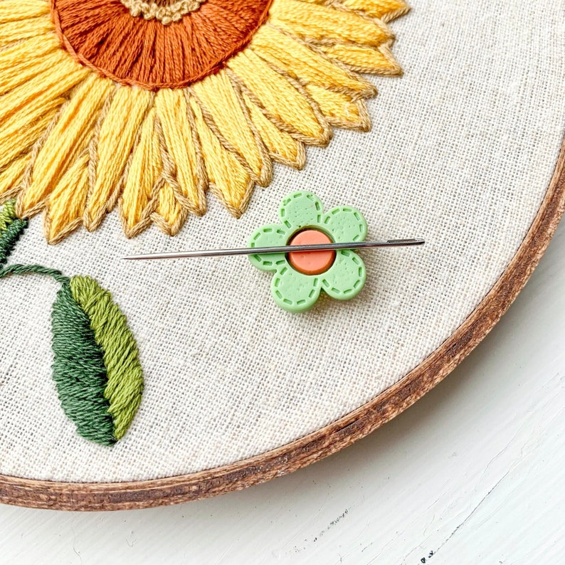 Green flower needle minder magnet holding an embroidery needle on embroidery hoop.