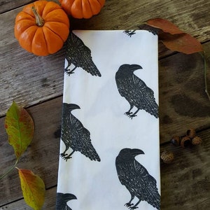 May include: White kitchen towel with a repeating black raven pattern. Two small orange pumpkins and fall leaves are arranged on a rustic wooden surface, creating a seasonal autumn theme.